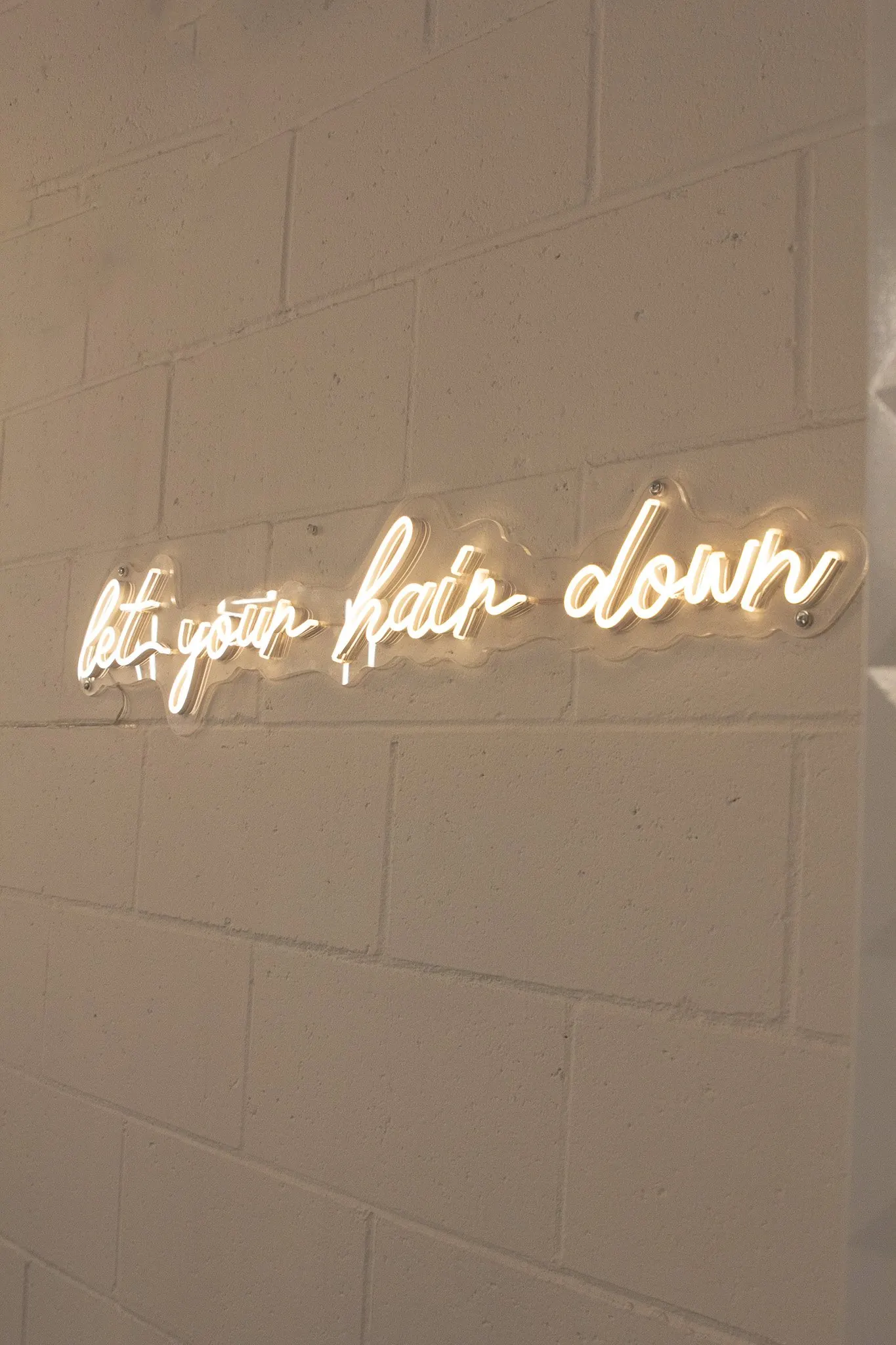 Neon script reading let your hair down on a white brick wall.